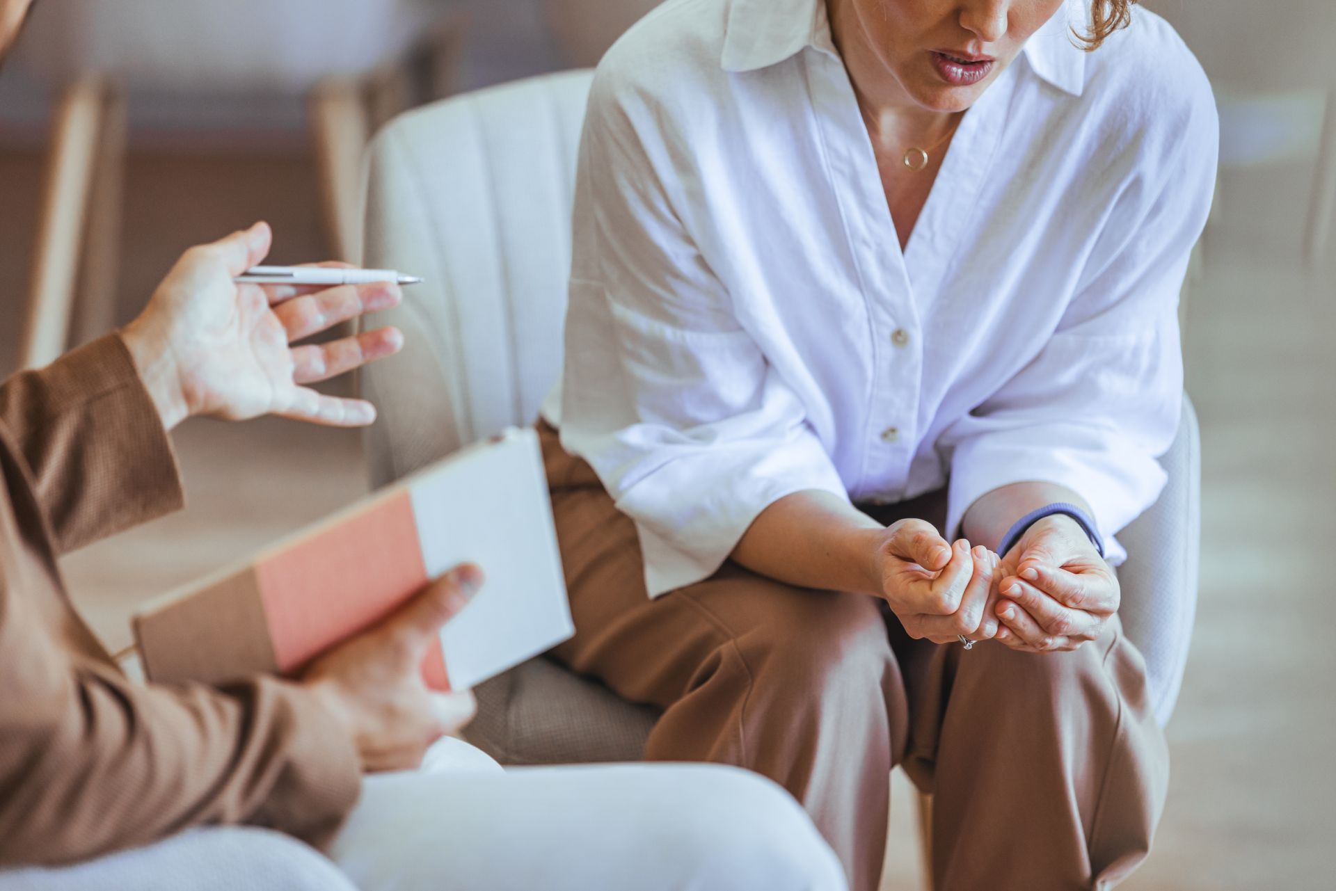 Person in white shirt seated on a couch during a conversation, holding hands clasped near their knees.