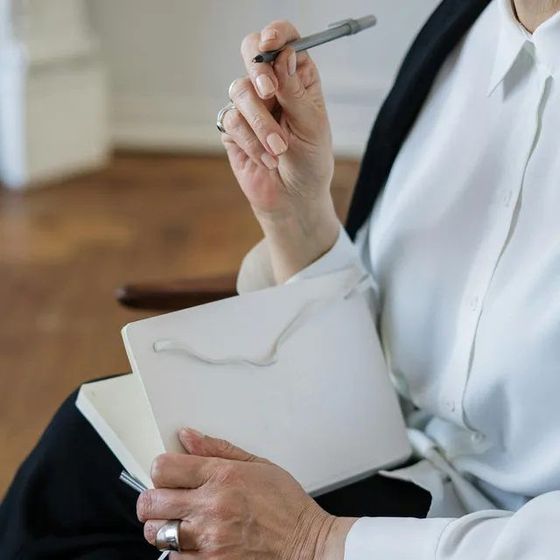Person in a white coat holding a pen and notebook, seated indoors.