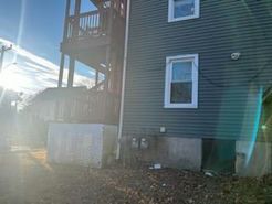 Side view of a house with dark siding, a deck, and concrete foundation. Overcast with sunlight.