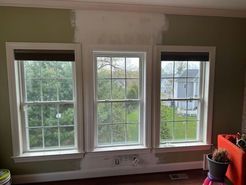 Three white-framed windows with dark shades; part of the wall painted white against green; brown floor and outside views.