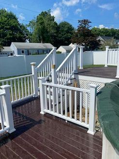White-railed deck with stairs, dark brown decking. Surrounding white fence, blue sky, and trees.