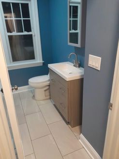Small bathroom with blue walls, wood-look vanity, white sink, toilet, and window with white trim.