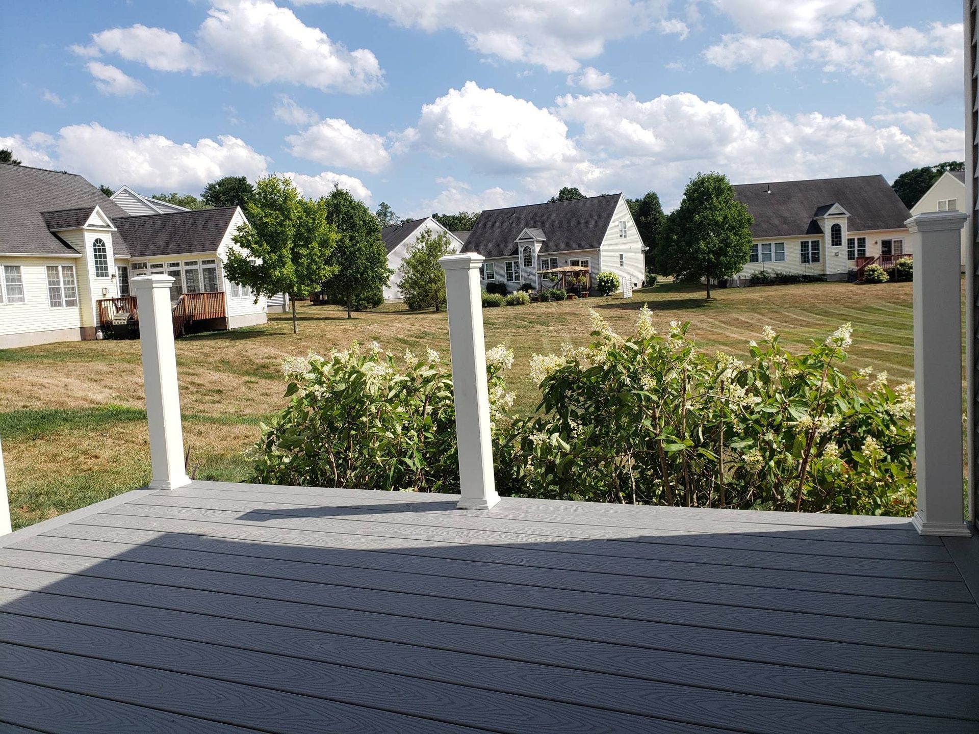 View from a gray deck overlooking a yard with houses and trees under a blue sky with clouds.