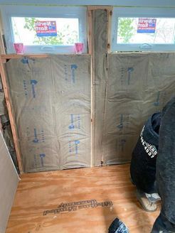 Interior construction with insulation between window frames; person crouches on wood floor.