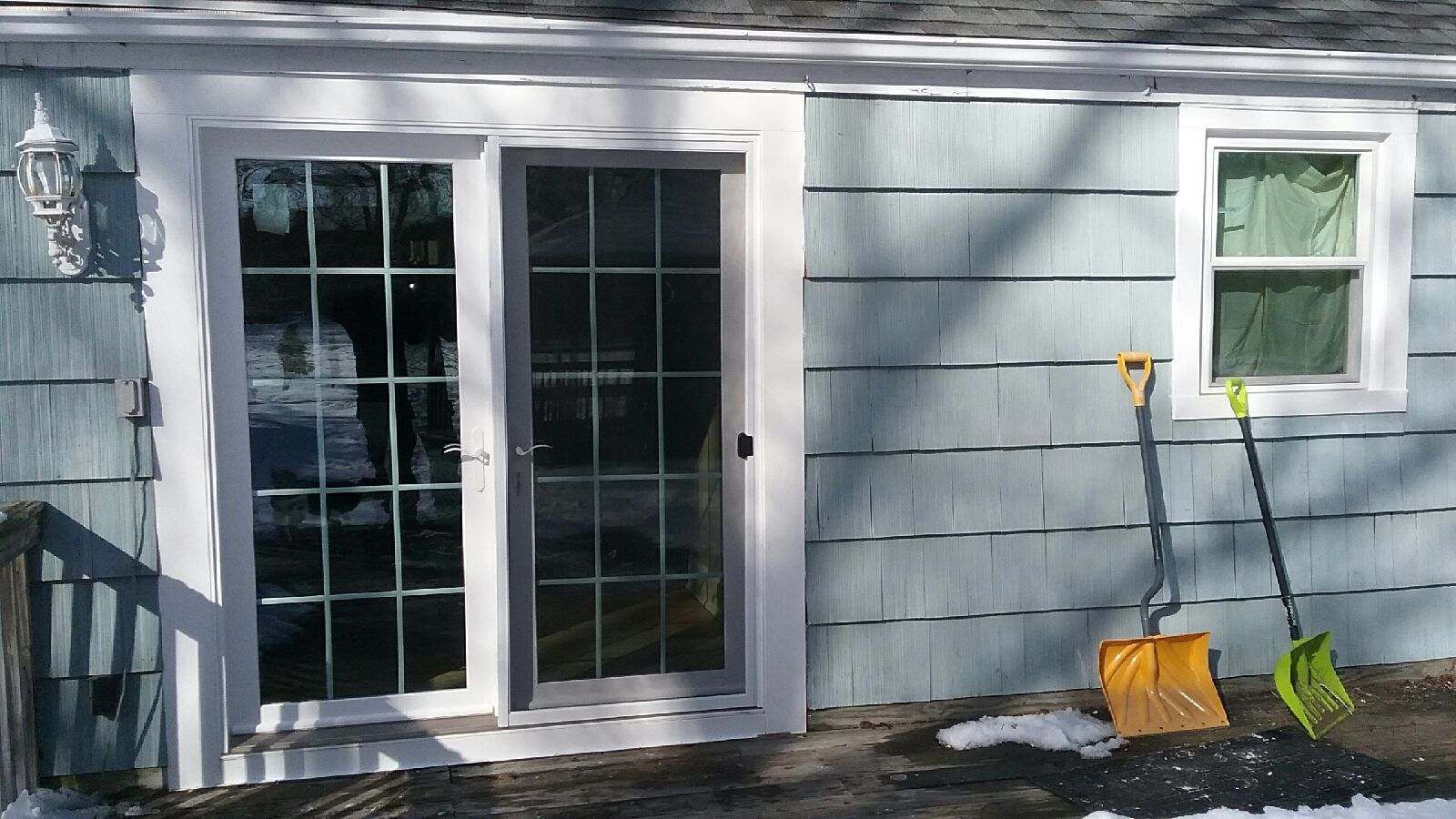 Sliding glass doors and window on a blue-shingled house; snow shovels lean against the wall.
