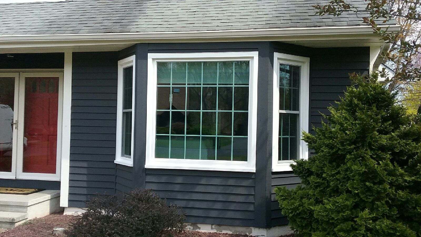 Dark gray house with white trim and a bay window. Red door and green bush.