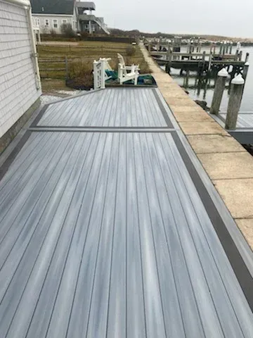 Gray wood deck next to a dock and water, with a white building in the background.