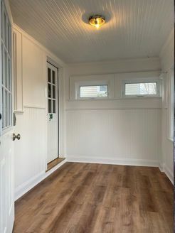 A small, bright room with white walls, wood-look floor, windows, and a door; ceiling light.
