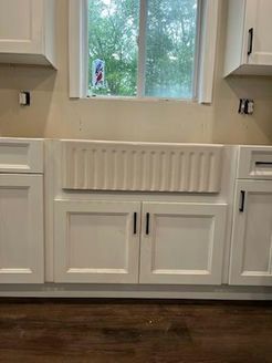 White farmhouse sink in kitchen cabinets beneath a window.