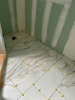 Bathroom floor tile installation in progress; white marble-look tiles with yellow spacers, unfinished green walls.