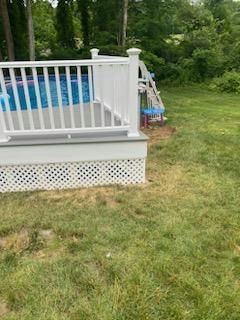 White deck with railing surrounds a blue above-ground pool. Lattice skirt. Grassy yard.