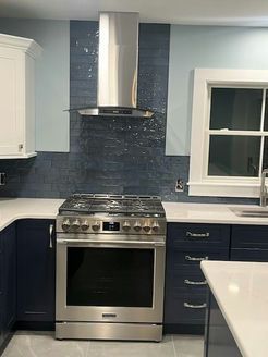 Kitchen with blue cabinets, stainless steel range, and range hood. Dark blue backsplash, white countertops, and window.