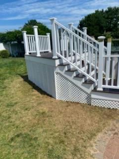 White deck with stairs, lattice skirting, and railings. Located on a grassy area, blue sky.