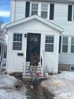 Person shoveling snow from steps of a white house with a porch.