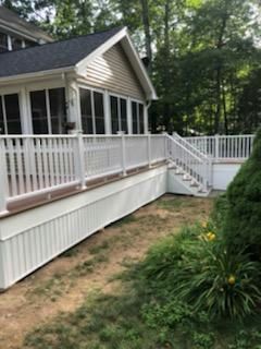 White deck with railing and stairs, attached to a house with screened porch, surrounded by grass and trees.