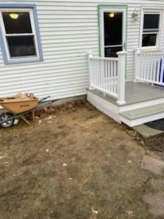 Wheelbarrow next to an empty dirt patch by a porch with white railings and a door, attached to a white house.