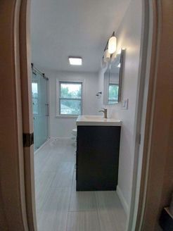 Bathroom interior through doorway: gray tile floor, dark vanity, glass shower, window, and bright overhead lights.