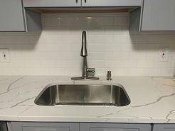 Stainless steel kitchen sink with faucet and soap dispenser on white countertop with gray cabinets and white tile backsplash.