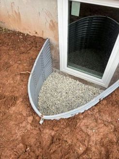Window well filled with gravel next to a window in a red dirt setting.