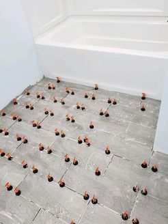 Bathroom floor tile installation in progress; gray tiles with leveling system visible near a white bathtub.