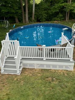 Above-ground swimming pool with a white deck and stairs, in a grassy backyard, surrounded by trees.