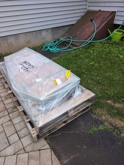 A pallet with a wrapped rectangular item on it sits outside a house, near a basement door.