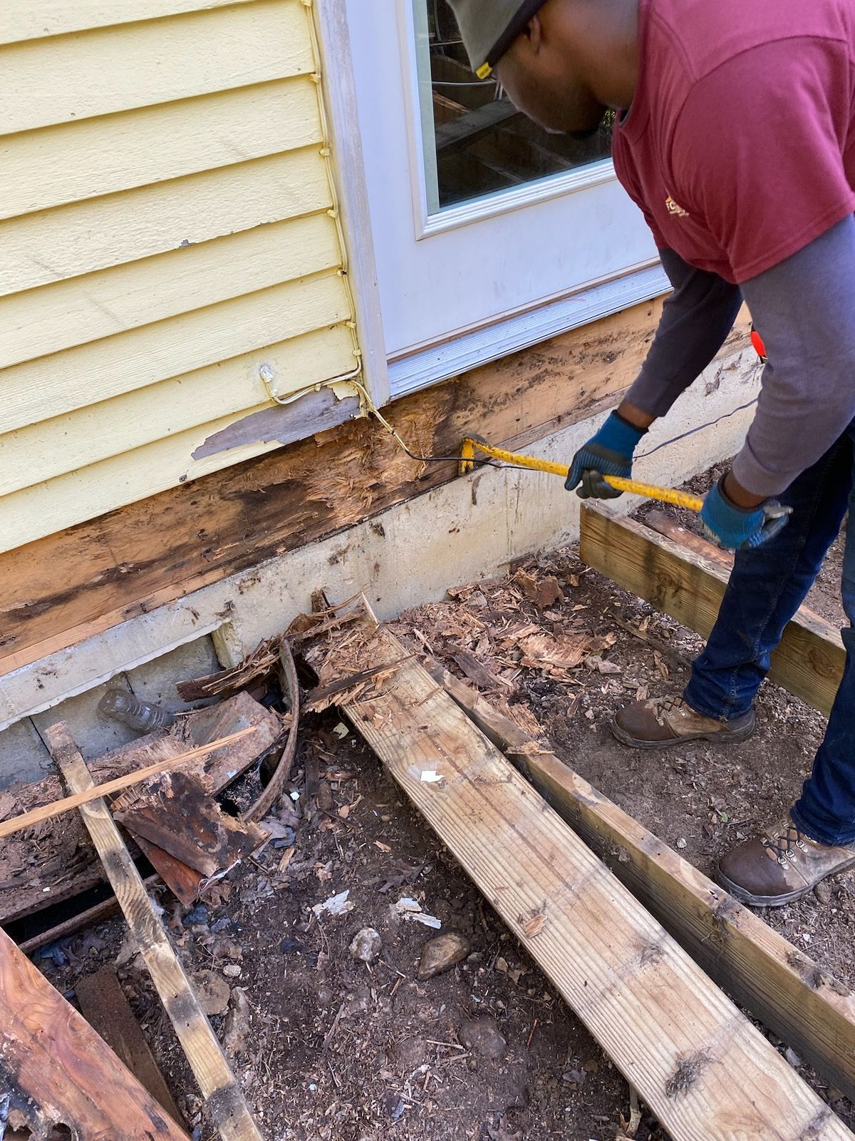 Man removing damaged wood siding from a house near a door, using a pry bar. Yellow siding, exposed wood.