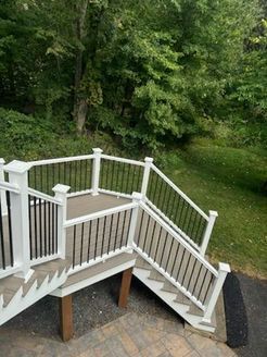 A multi-level deck with white railings, black balusters, and brown wood supports, set against a green wooded background.
