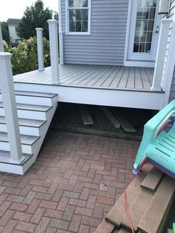 Gray deck and stairs with white posts, built over brick patio. A turquoise chair sits nearby.