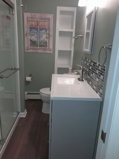 Bathroom with gray vanity, white countertop, tall storage, shower, and window art; muted green walls, dark wood-look floor.