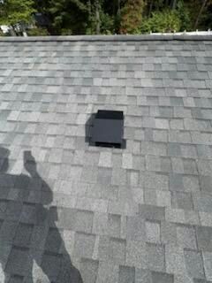 A dark vent box on a gray shingle roof. The shadow of a person holding a phone is visible.