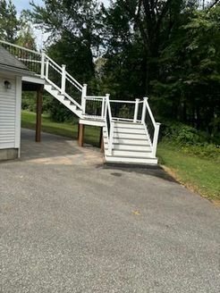 White staircase descends from a white-sided building with railings, leading to a dark asphalt driveway. Green trees in background.