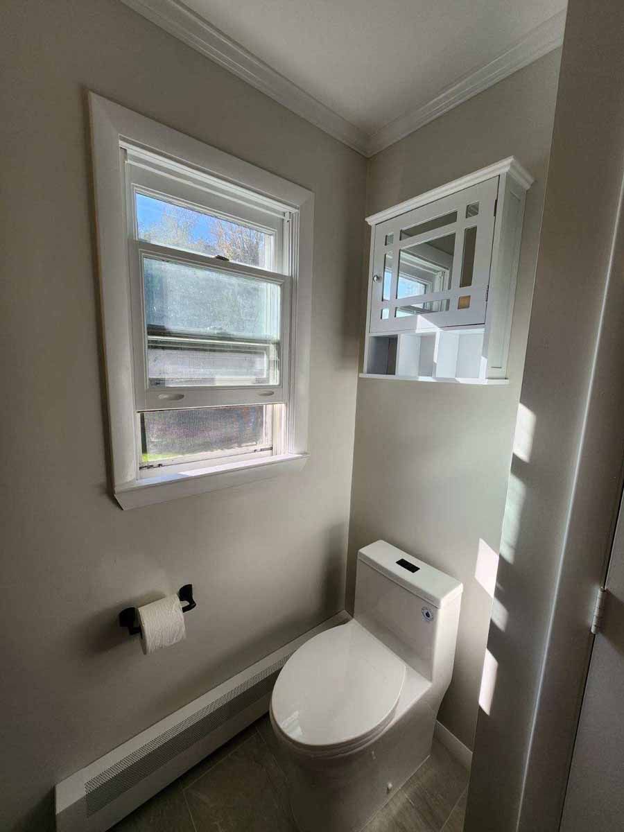 A small bathroom with a toilet, window, and wall-mounted cabinet. The walls are light grey.