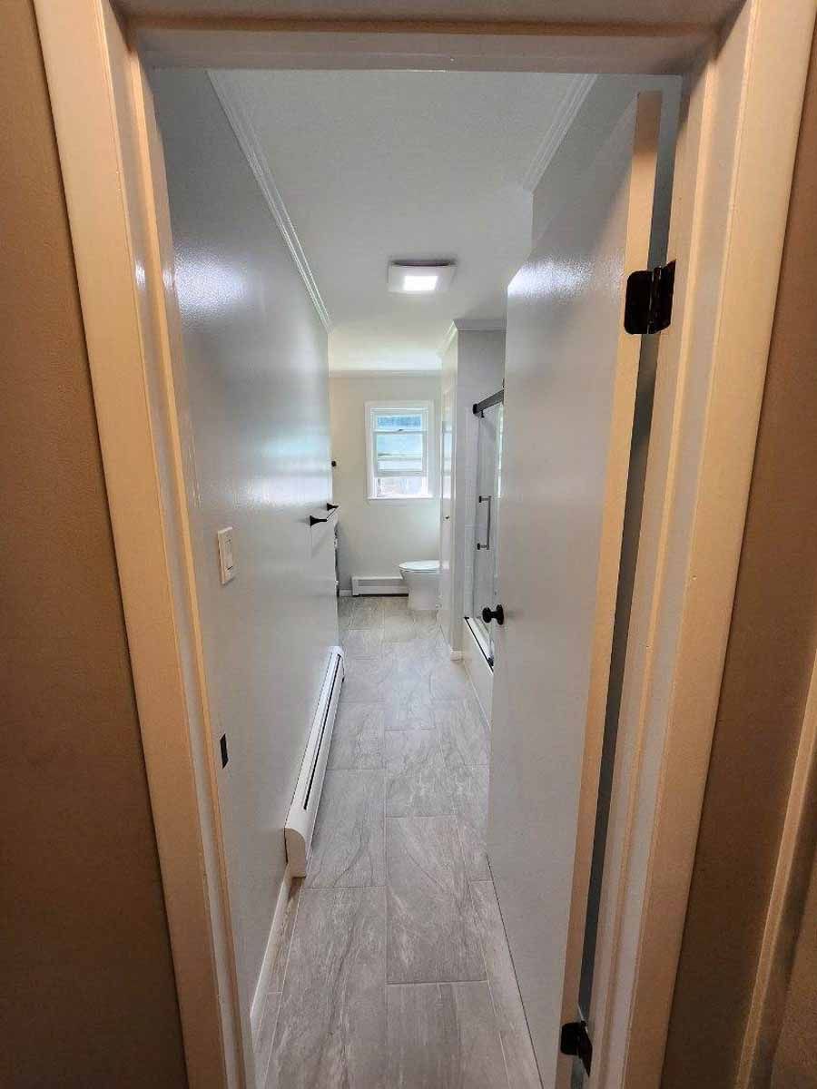 Narrow hallway with light gray flooring and walls, leading to a bathroom with a window.