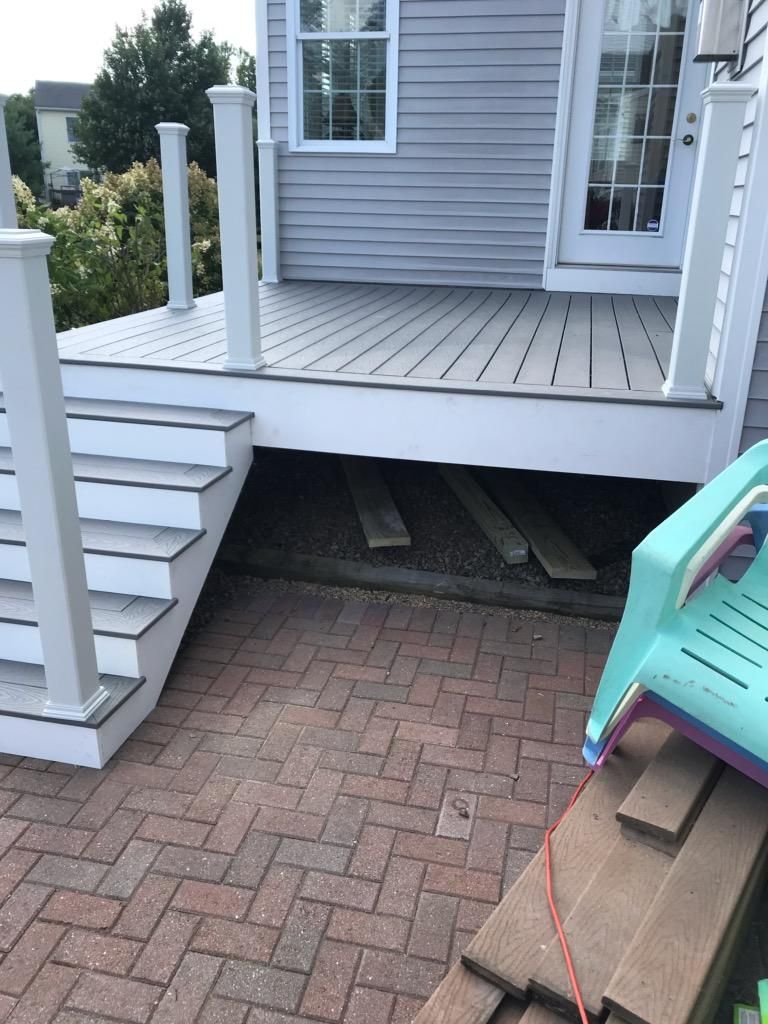 Gray deck with stairs leading down to a brick patio. A plastic chair and wood scraps are present.