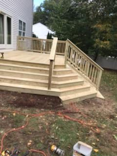 Wooden deck with stairs, built next to a white house, surrounded by grass and trees.