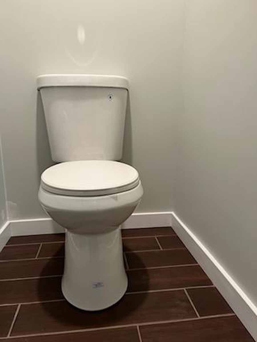 White toilet in a small bathroom corner. Brown tile floor, white baseboards, and gray walls.