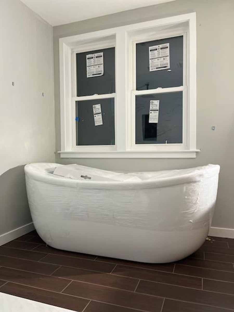 White bathtub wrapped in plastic under a window in a bathroom with dark brown floor tiles.