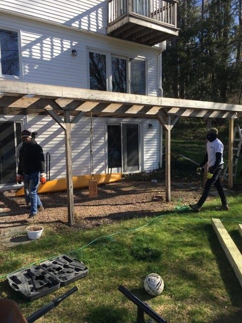 Two people working on a deck project outside a white house. One saws wood, other walks towards them.
