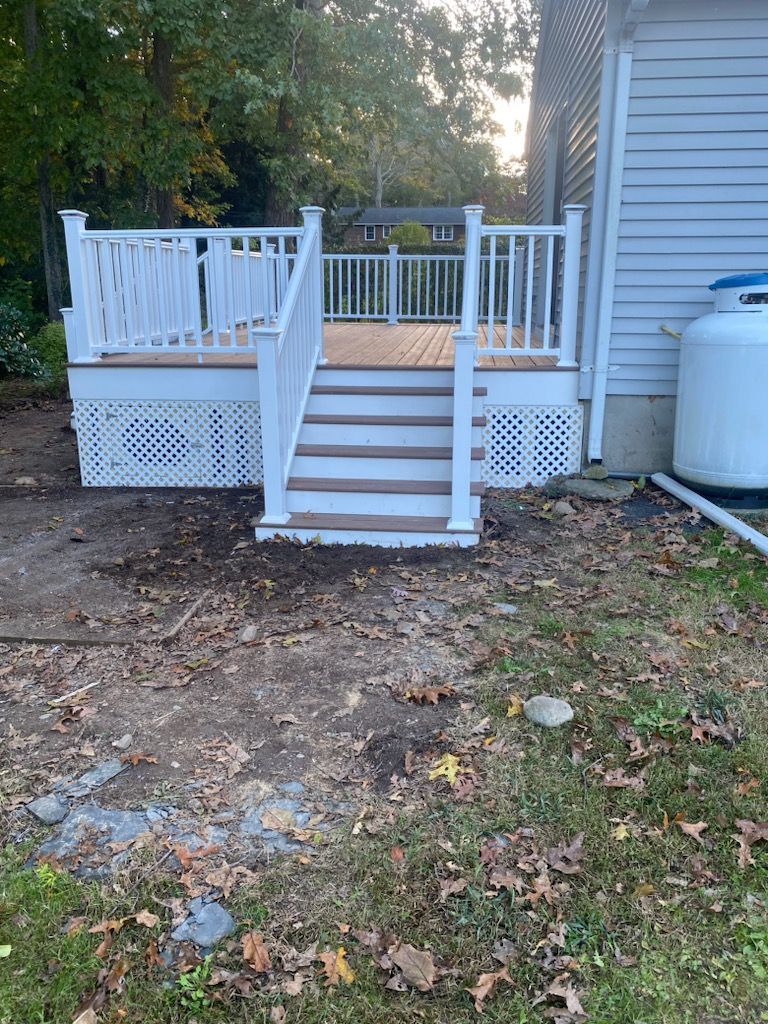 White deck with lattice skirting, stairs, and railing, attached to a gray house with a propane tank.