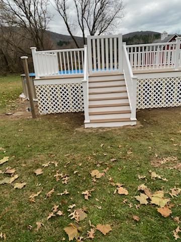 White deck with stairs leading to above-ground pool; latticework, grass, and trees are in the foreground.