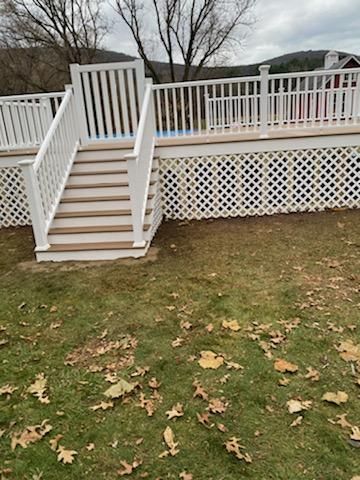 White deck with stairs, lattice skirting, and railing, on grass covered in fallen leaves, with trees in background.