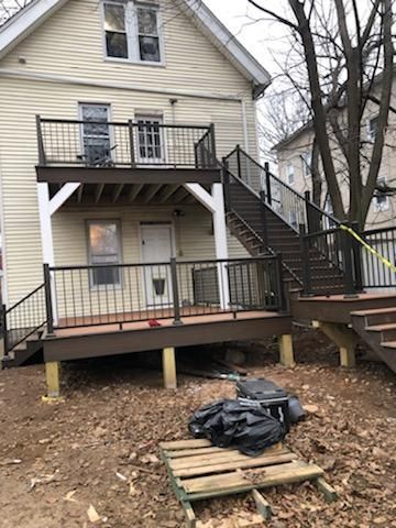 Two-story house with decks and stairs. Brown decks, black railings, and tan siding. Unfinished backyard.