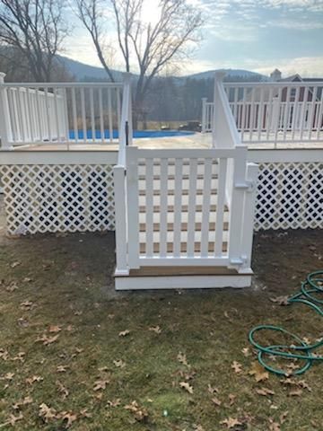 White gate on deck leads to a swimming pool. Lattice detailing and railing surround the deck. Green grass.