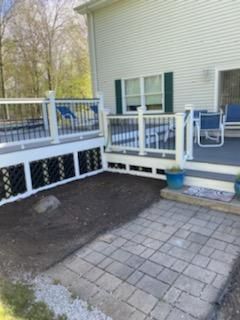 Deck with white railing and black accents, adjacent to a house with gray pavers and a dirt patch.