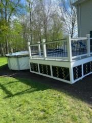 Deck with white railings and black accents next to a spa. Green grass and trees in the background.
