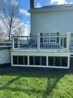 White deck with black railings and decorative lattice panels, attached to a light-colored house, on a green lawn.