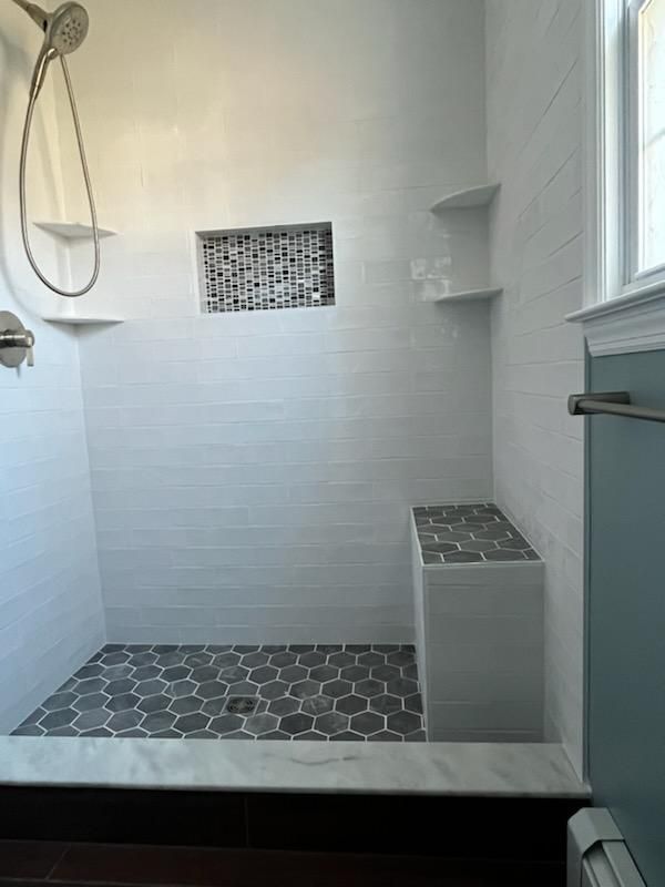 Bathroom shower with white and grey tile, built-in bench, shelves, and a dark tile floor.