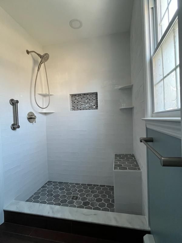 Modern bathroom shower with white tiled walls, gray and white mosaic floor, built-in bench, and a window.