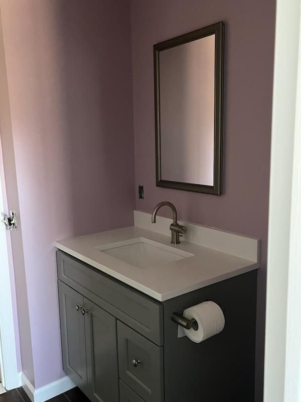 Gray bathroom vanity with white countertop, faucet, and mirror. Purple wall.